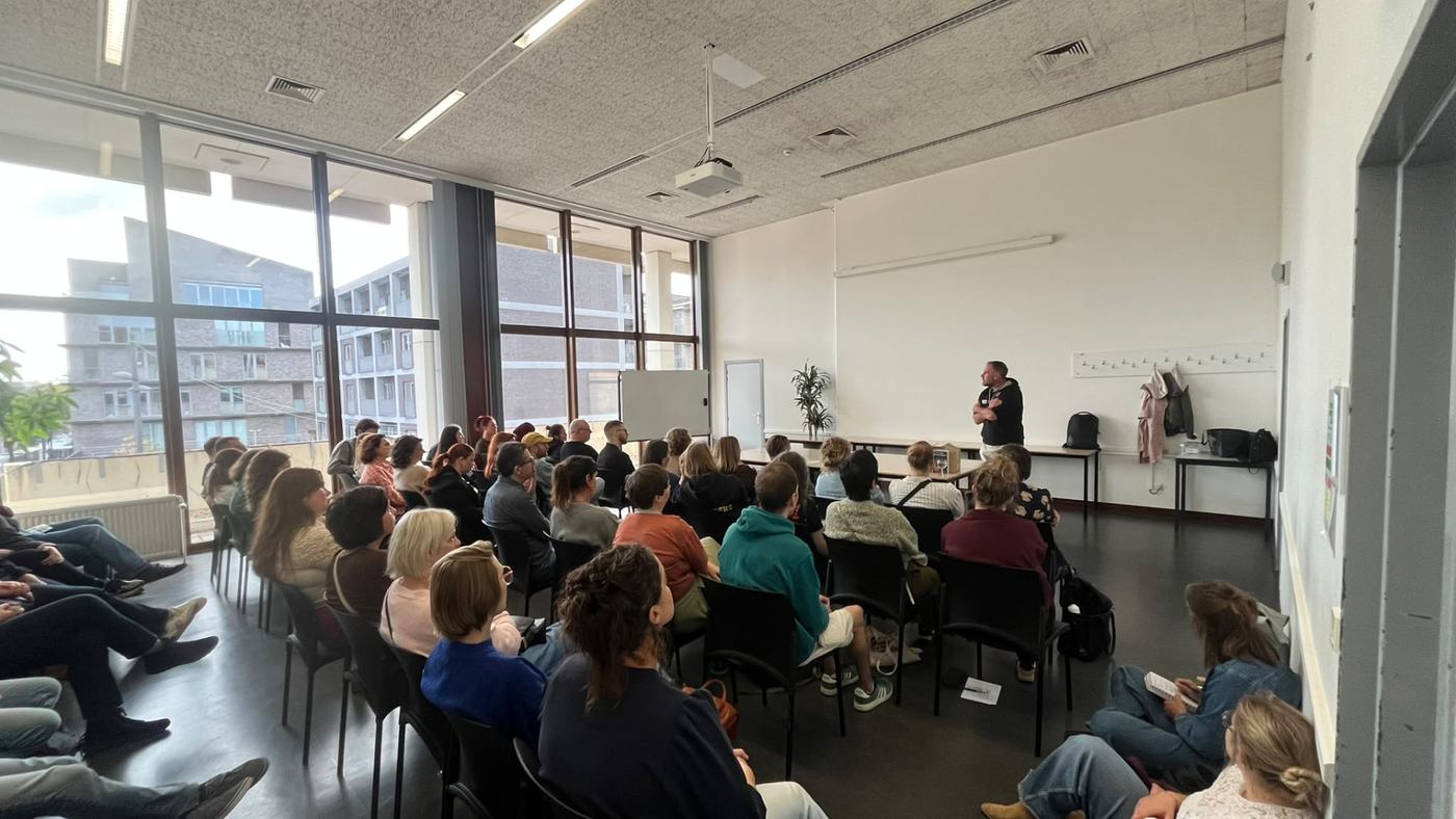 Kenneth staat met gekruiste armen vooraan een lokaal en spreekt een groep mensen toe