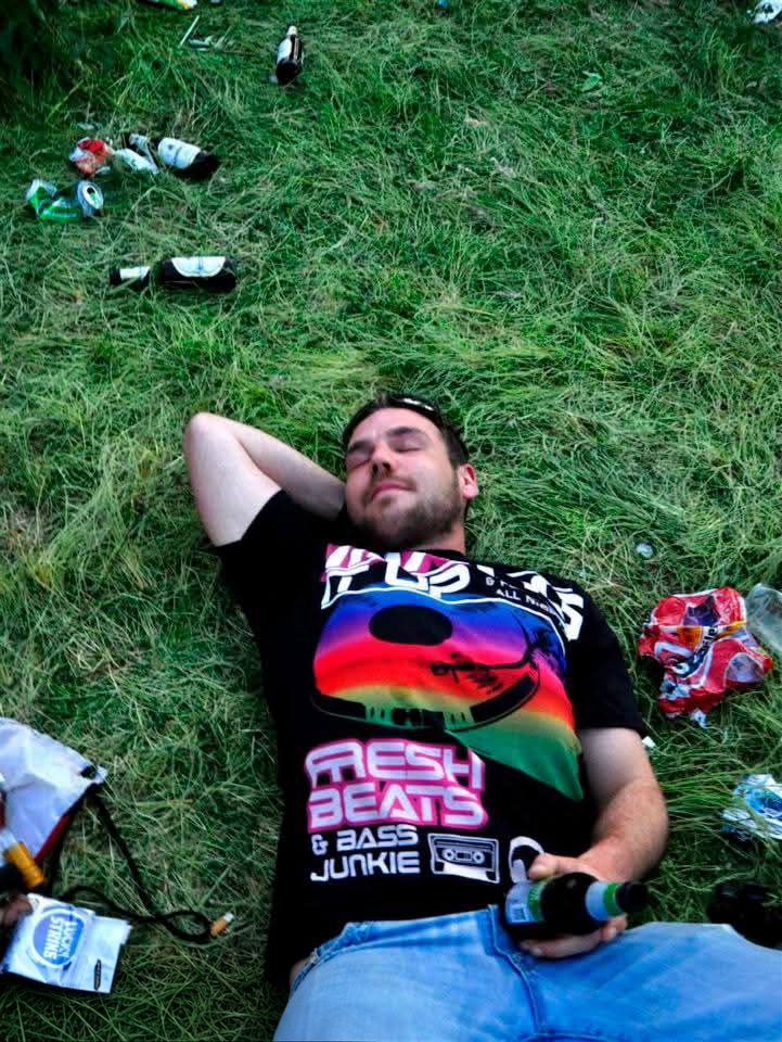 Kenneth ligt met gesloten ogen in het gras met een flesje in zijn hand, omringd door lege flessen en blikjes